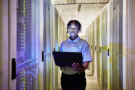 Medium-shot-portrait-of-female-computer-engineer-holding-laptop-while-working-in-row-of-servers-in-data-center_GettyimagesThomas-Barwick