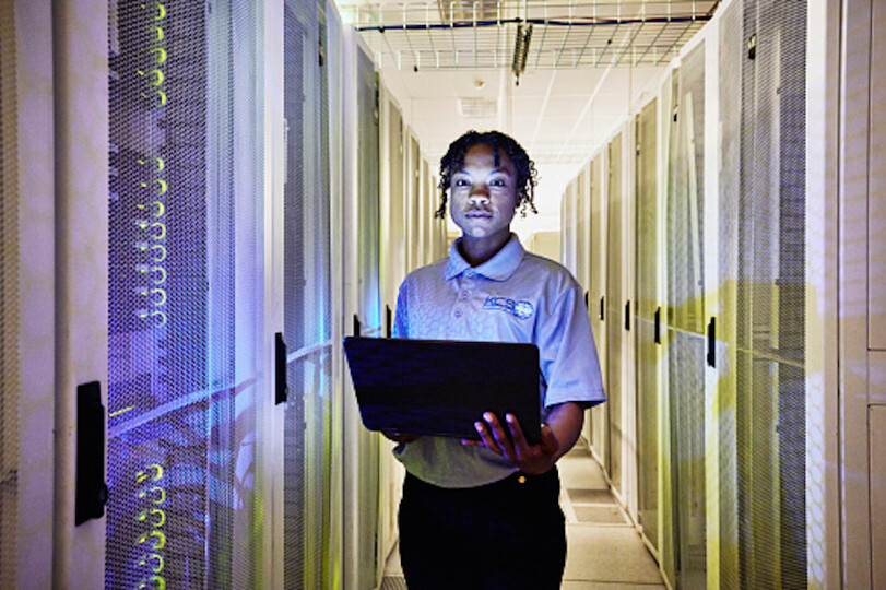 Medium-shot-portrait-of-female-computer-engineer-holding-laptop-while-working-in-row-of-servers-in-data-center_GettyimagesThomas-Barwick